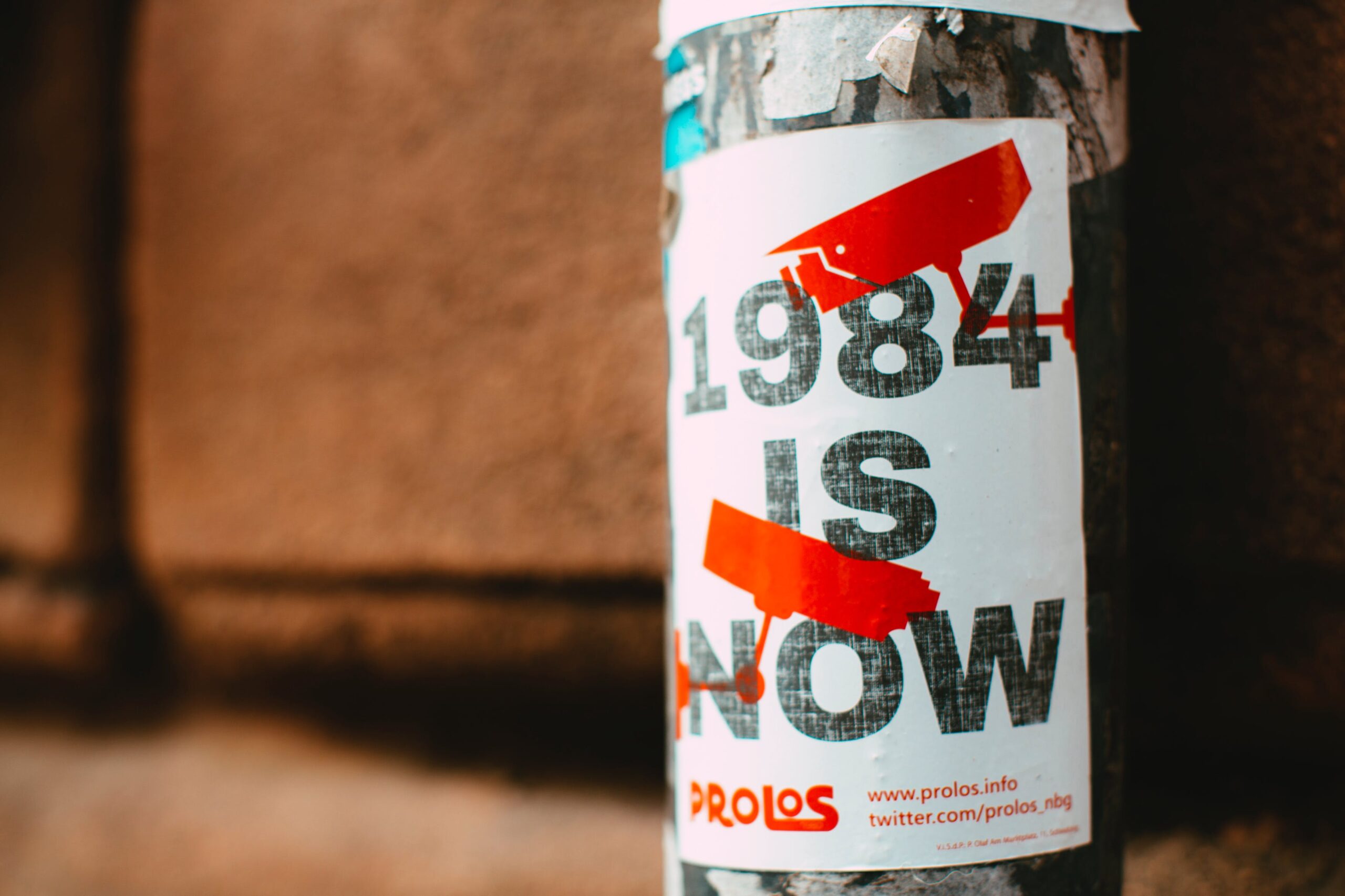 A well-worn poster, stuck to a streetlight or telephone pole, stating "1984 IS NOW"